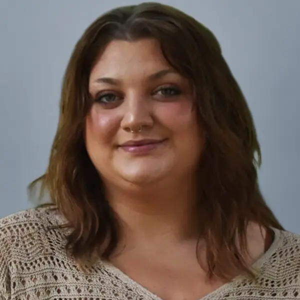 Smiling woman with shoulder-length brown hair wearing a light knit top against a gray background