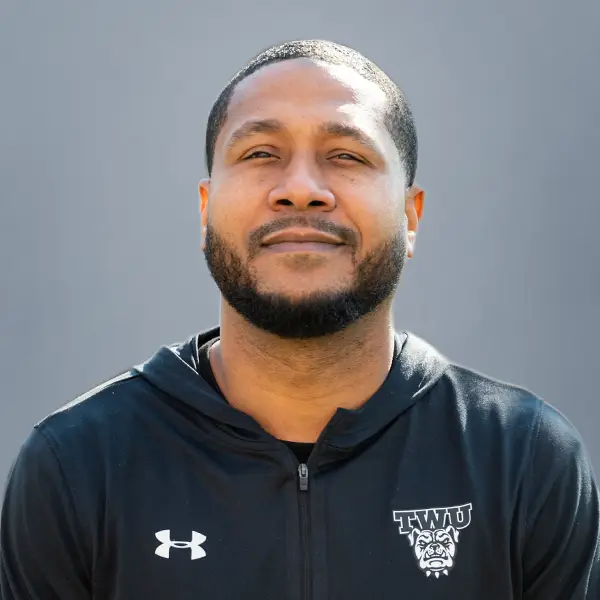 Portrait of a man with a short beard and mustache, wearing a black zip-up hoodie with an Under Armour logo and a TWU bulldog emblem on the chest.