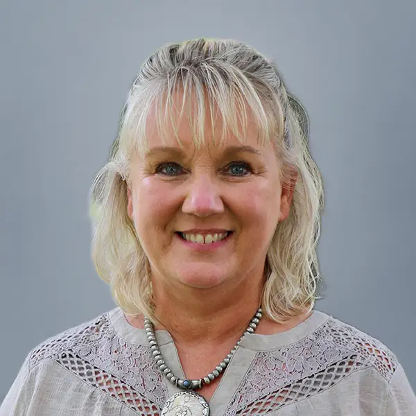Smiling woman with blonde hair and bangs, wearing a light lace top and beaded necklace, posing outdoors against a gray background.