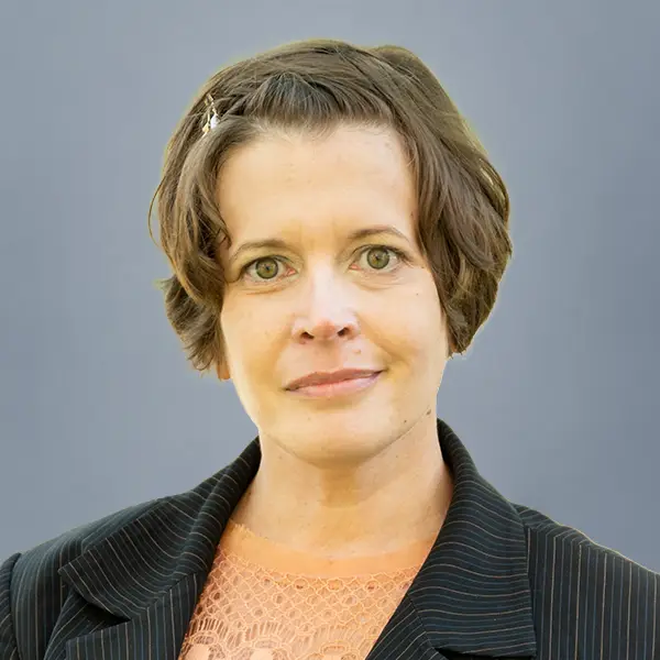 Head-and-shoulders portrait of a woman with short brown hair, wearing a pinstripe blazer and peach lace top against a gray backdrop.
