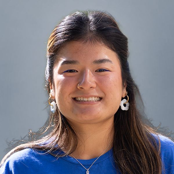 Portrait of a smiling Asian woman with long dark hair, wearing a blue top and white flower-shaped earrings, against a gray background, centered in the frame.