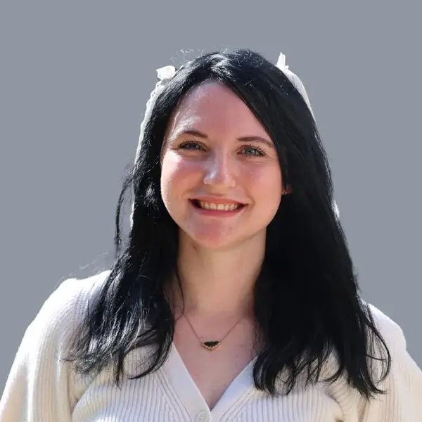 Portrait of a smiling woman with long dark hair, wearing a white cardigan and a small pendant necklace, against a gray background.