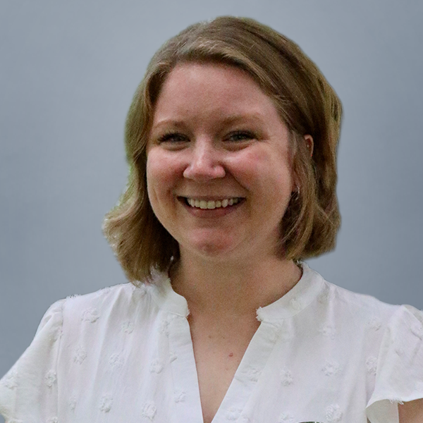 Smiling woman with shoulder-length blonde hair wearing a white blouse against a gray-blue background.