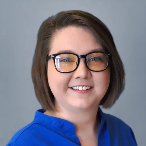 Portrait of a smiling woman with short brown hair, black glasses, and a blue blouse against a gray background.