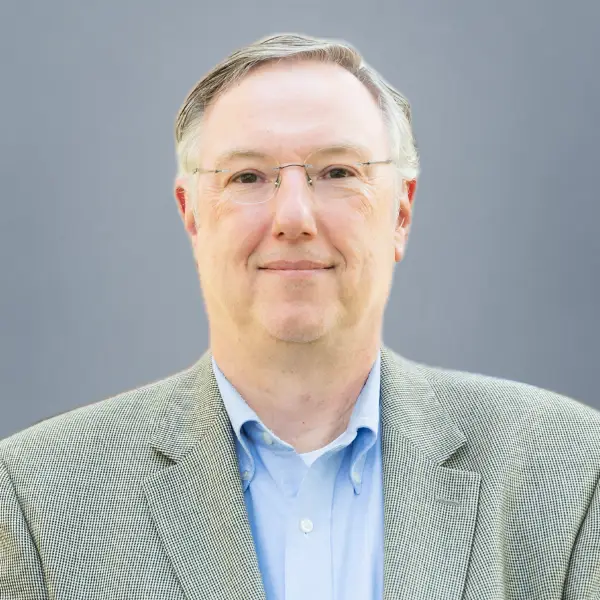 Head-and-shoulders portrait of a smiling middle-aged man with glasses, wearing a light blue shirt and gray blazer against a gray backdrop.