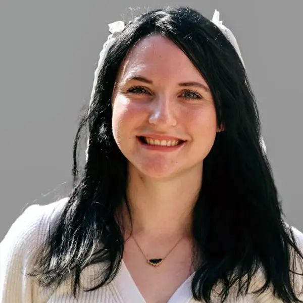 Smiling woman with long dark hair, wearing a white sweater and a small pendant necklace, neutral background.