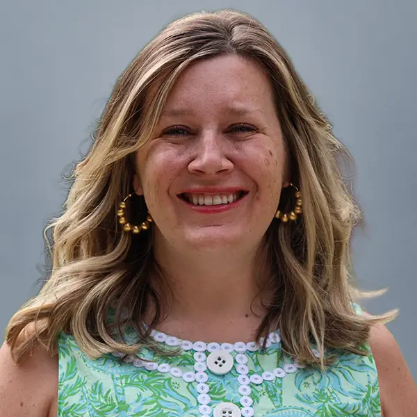 Smiling woman with shoulder-length blonde hair, wearing a green patterned dress and gold hoop earrings against a gray backdrop.