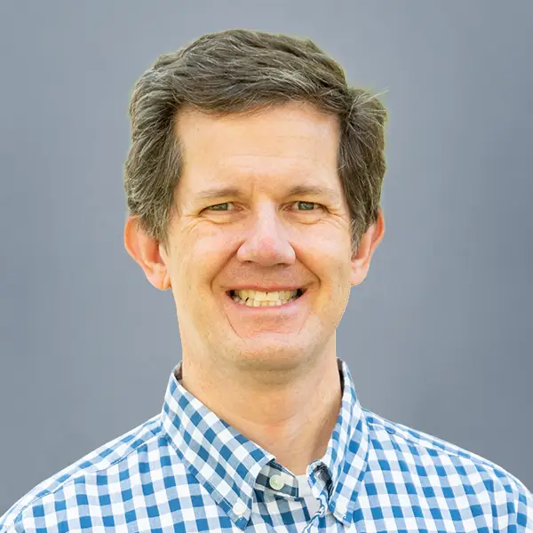 Smiling man in a blue-and-white checkered shirt, facing the camera, against a gray background.
