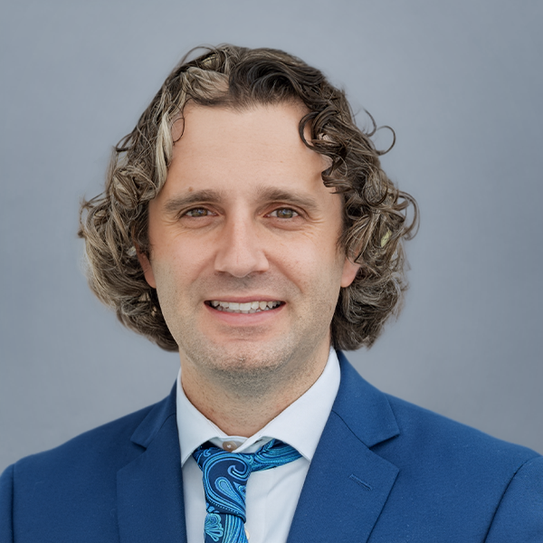 Headshot of a smiling man in a blue suit and patterned blue tie against a gray background for a corporate profile.