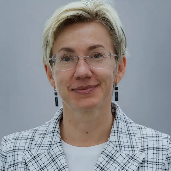 Professional headshot of a woman with short blonde hair and glasses, wearing a white plaid blazer, facing the camera.