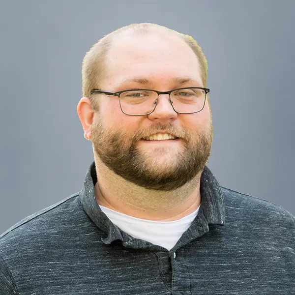 Smiling man with glasses and beard, wearing a dark collared shirt, against a gray background (head-and-shoulders portrait).