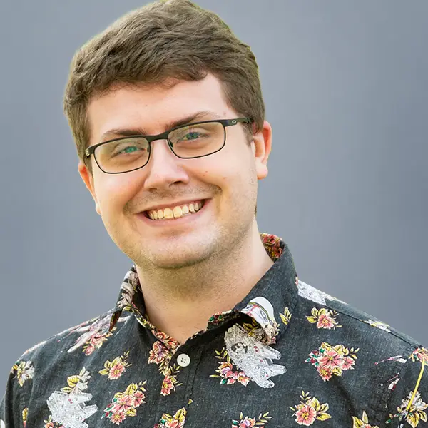 Smiling man with short brown hair and glasses wearing a dark floral button-down shirt against a gray background.