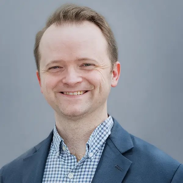 Smiling man in a blue blazer and checkered shirt posing for a professional headshot against a gray background.
