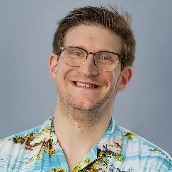 Smiling man with glasses wearing a tropical Hawaiian shirt against a light blue background.