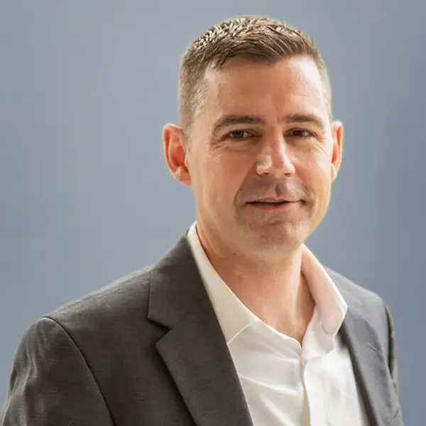Professional headshot of a man in a dark gray suit and white shirt against a blue background.