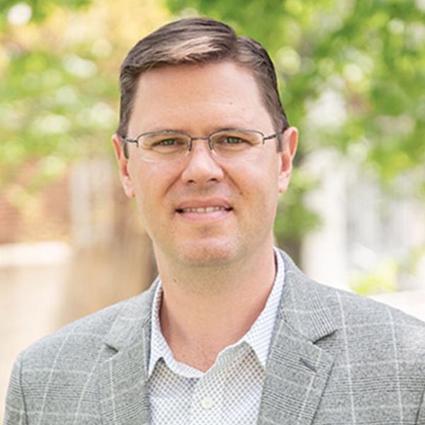 Professional headshot of a man wearing glasses and a gray plaid blazer outdoors with greenery in the background