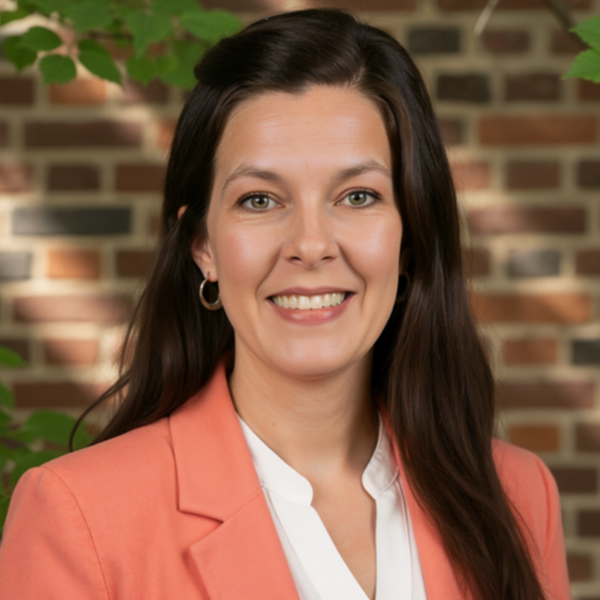 Professional headshot of a smiling woman with long dark hair, wearing a coral blazer and white blouse, with a brick wall and greenery in the background.