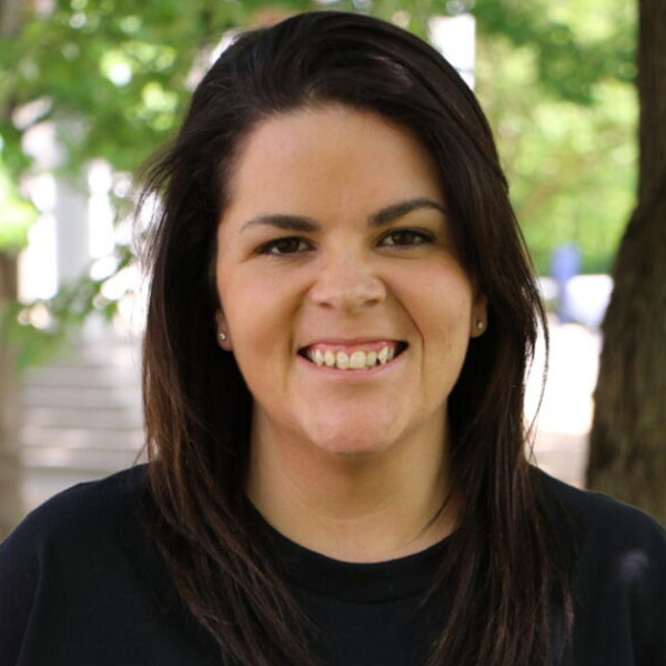 Smiling woman with dark hair in a black shirt, outdoors with trees in the background.