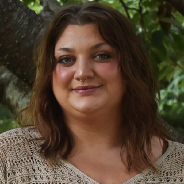 Portrait of a smiling woman with shoulder-length brown hair wearing a beige crochet top, outdoors with greenery in the background.