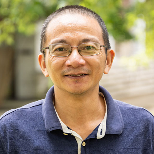 Portrait of a smiling middle-aged man with glasses, wearing a navy polo shirt outdoors with greenery in the background.
