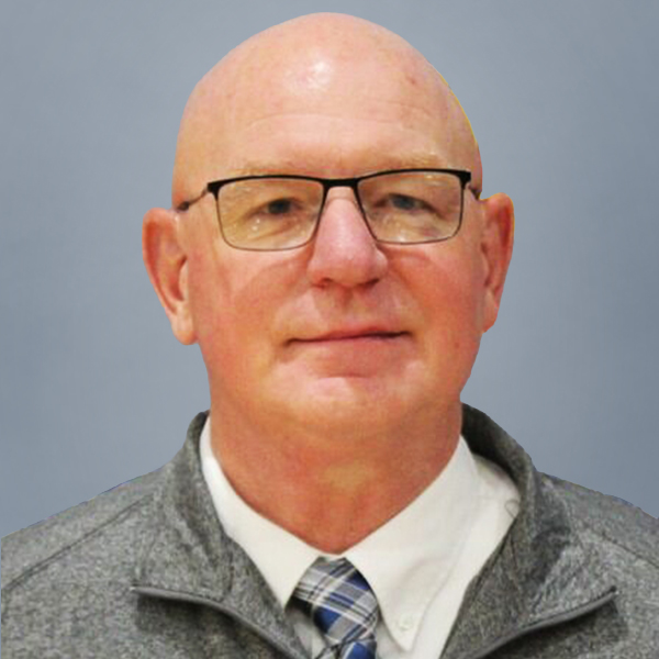Portrait of a bald man wearing glasses, white shirt, blue plaid tie, and gray jacket against a neutral blue background.