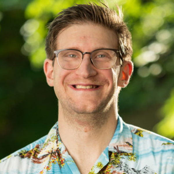 Smiling man with glasses wearing a tropical shirt outdoors, green foliage in the background.