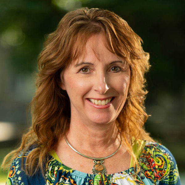 Smiling woman with wavy light brown hair outdoors, wearing a colorful patterned top and a silver necklace.