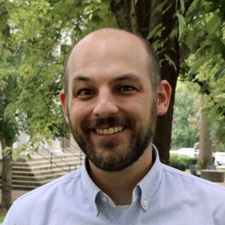 Smiling man with a beard outdoors, wearing a light blue check shirt, in a green park setting.