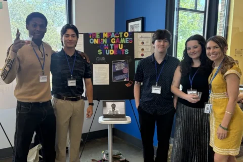 Six students with lanyards pose beside a research poster about the impact of online games on TWU students, with a laptop on a small table nearby.