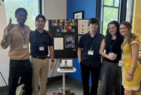 Six students with lanyards pose beside a research poster about the impact of online games on TWU students, with a laptop on a small table nearby.