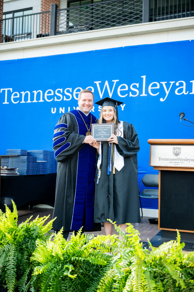 Graduate in cap and gown holding an award plaque with a faculty member in ceremonial robes on stage at Tennessee Wesleyan University, blue backdrop.