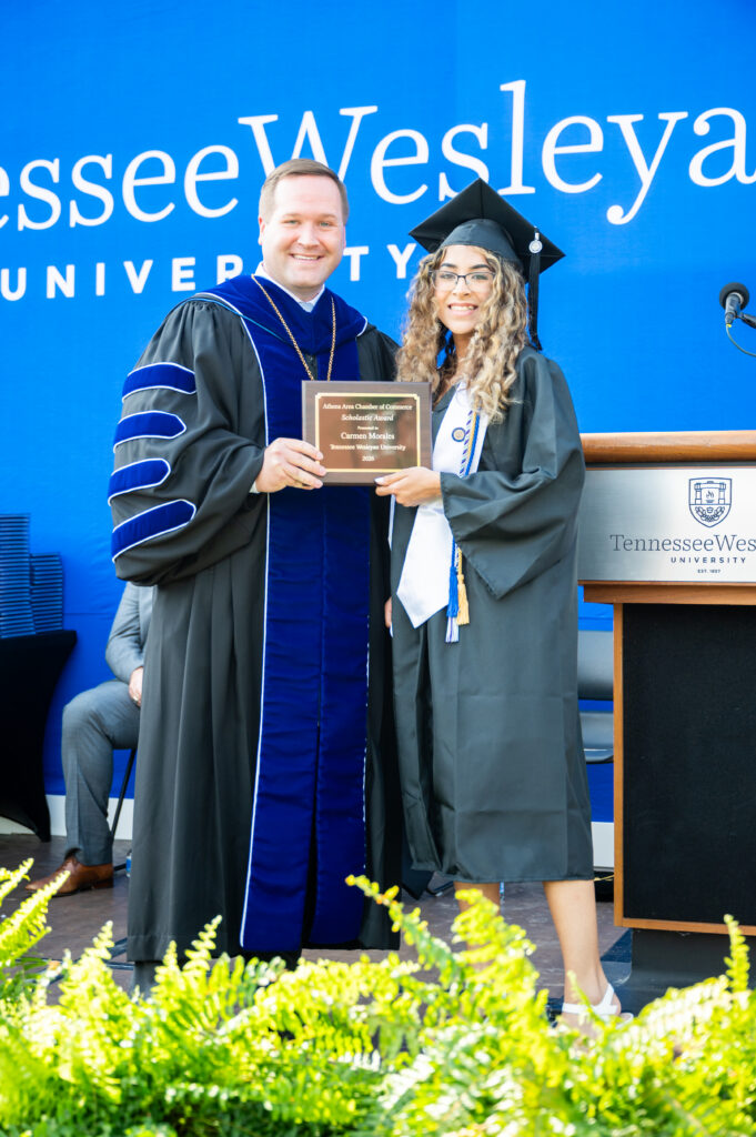 Faculty member in blue and black regalia presents a plaque to a graduate in cap and gown on stage with a Tennessee Wesleyan backdrop behind them.