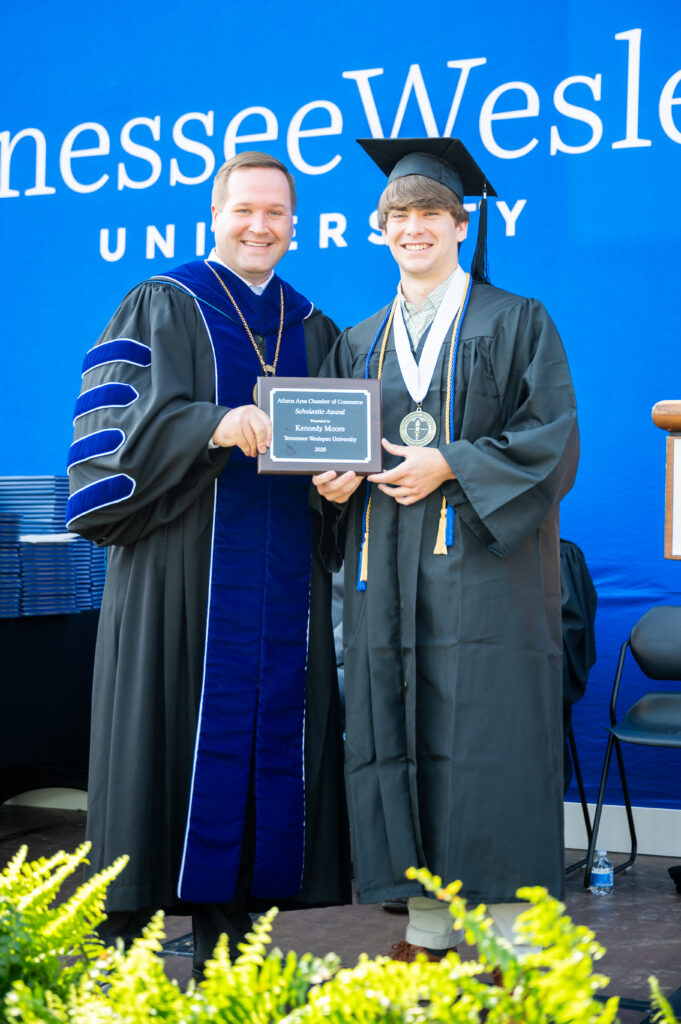 Two graduates pose for a photo in cap and gown, holding an award plaque during a university ceremony in front of a blue backdrop.