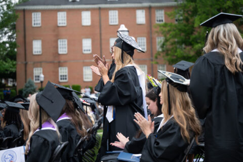 Graduates in black caps and gowns applaud during an outdoor commencement ceremony on campus.