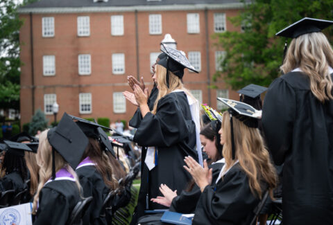 Graduates in black caps and gowns applaud during an outdoor commencement ceremony on campus.