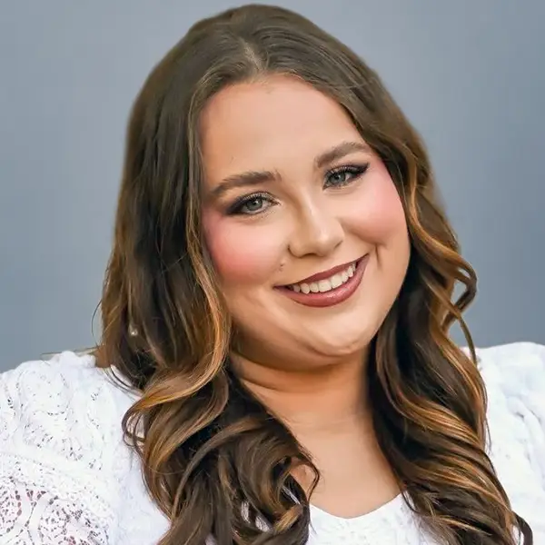 Smiling woman with long wavy brown hair and a white lace top, posed against a blue-gray background.
