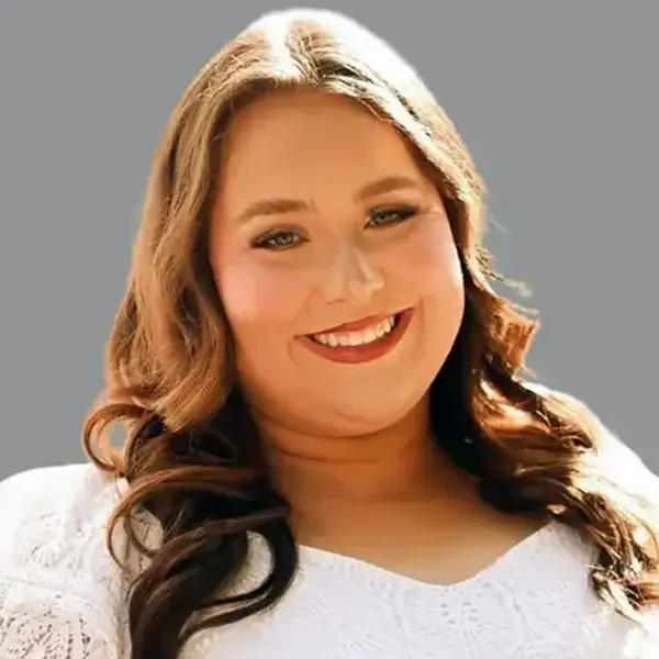 Portrait of a smiling woman with long wavy brown hair wearing a white lace top, against a gray background.