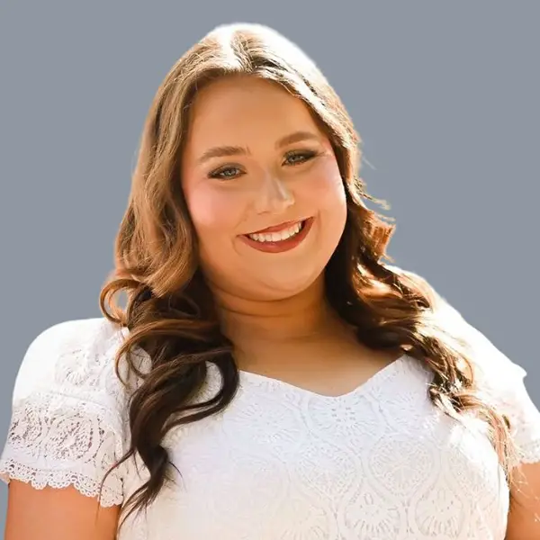 Smiling woman with long wavy hair wearing a white lace top, against a gray background.
