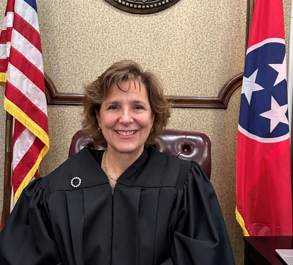 Female judge in a black robe seated at a desk, with the United States flag on her left and another flag on her right in a courtroom backdrop.