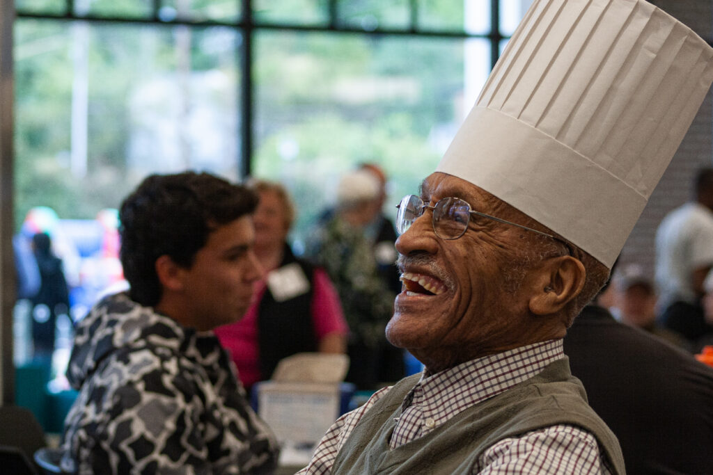 | Tennessee Wesleyan University Smiling elderly man wearing a tall white chef's hat and glasses at a social event.