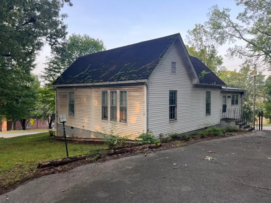 White siding house with a steep roof covered in ivy, surrounded by trees and a paved driveway.