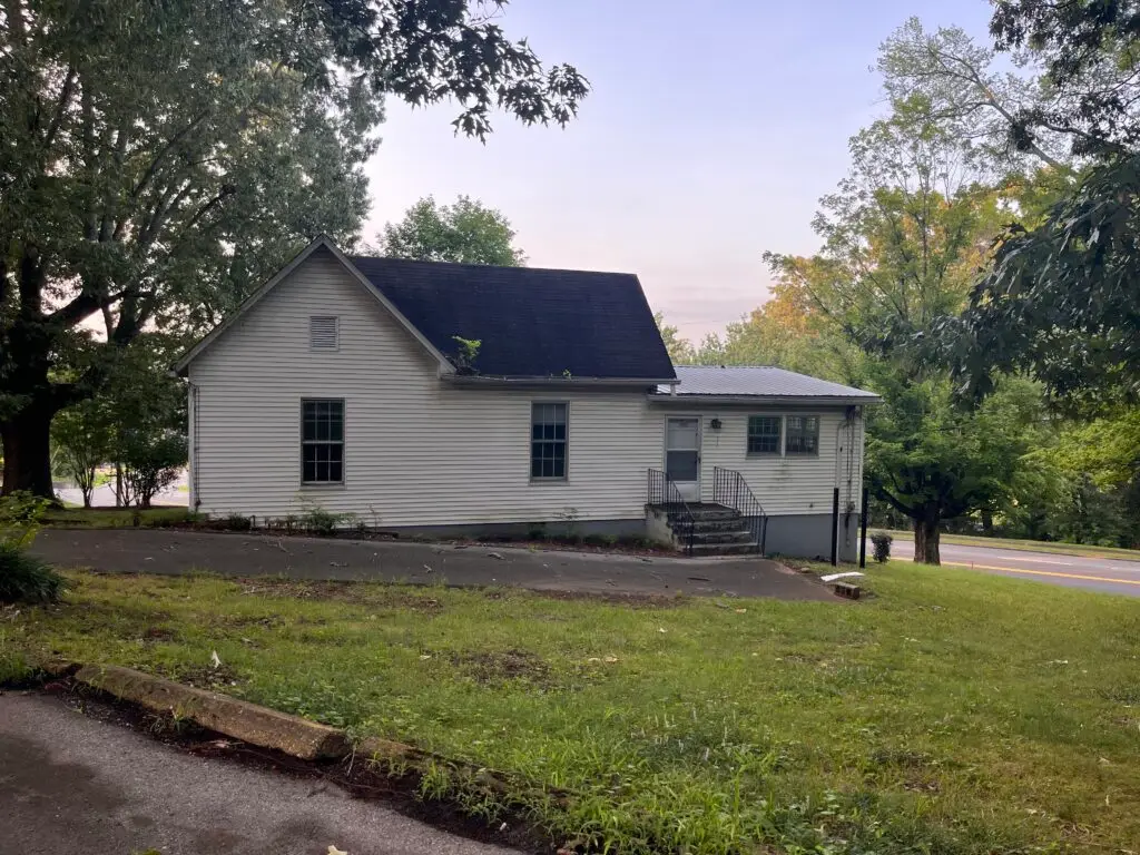 White house with a dark roof framed by tall trees, a paved driveway in front and a street visible to the right