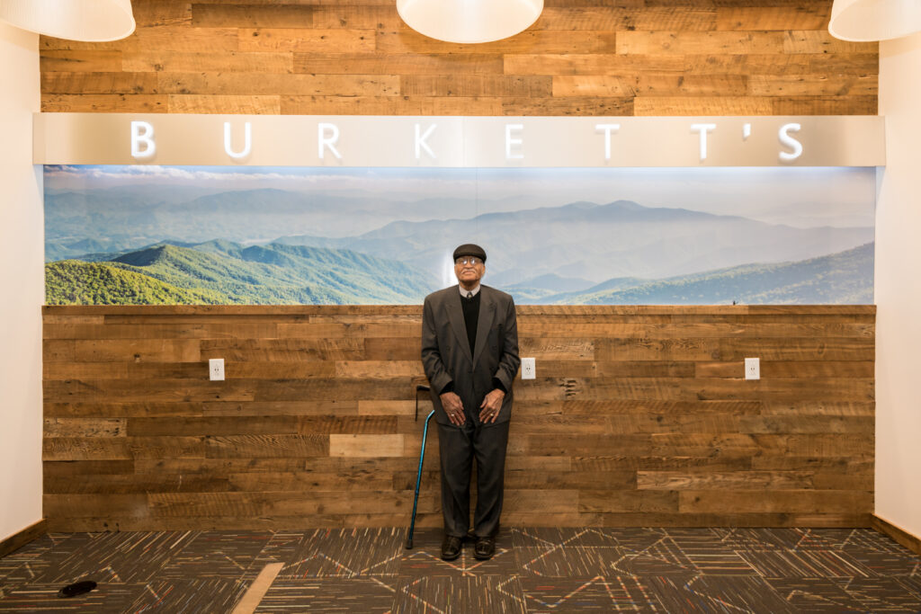 | Tennessee Wesleyan University Older man in a dark suit and flat cap stands with a cane before a wooden wall and mountain mural.