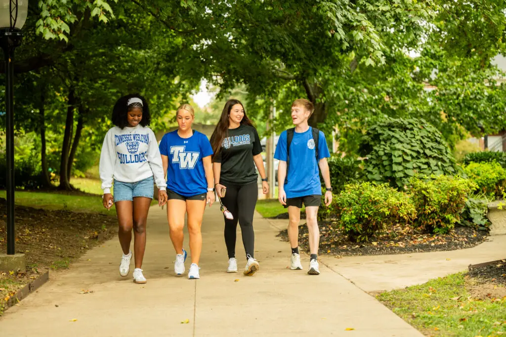 Four college students walk together on a sunny campus path surrounded by trees, wearing athletic tops.