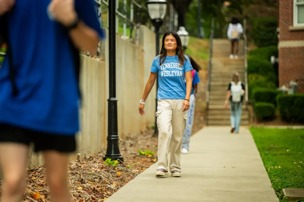 Young woman wearing a Tennessee Wesleyan t-shirt walks along a campus sidewalk, smiling.
