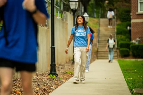 Young woman wearing a Tennessee Wesleyan t-shirt walks along a campus sidewalk, smiling.