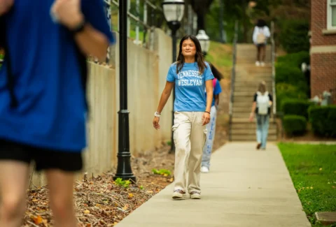 Young woman wearing a Tennessee Wesleyan t-shirt walks along a campus sidewalk, smiling.