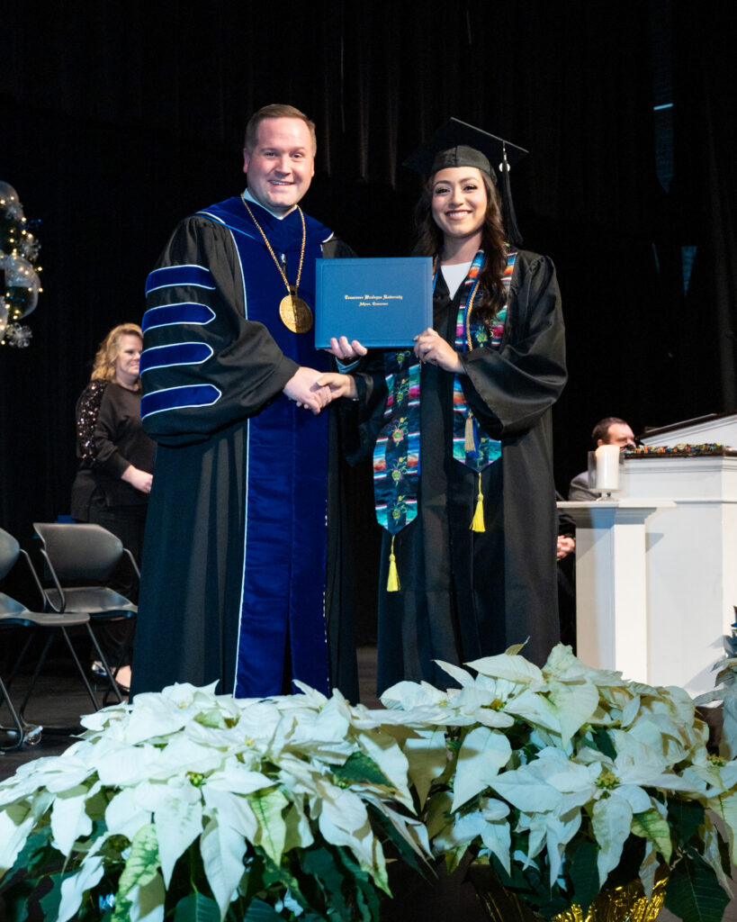 Graduate in cap and gown receives diploma from faculty member on stage, blue university regalia and a diploma folder visible