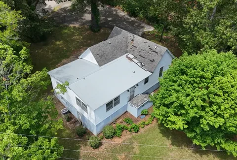 Aerial view of a white two-story house with a gray roof, surrounded by green trees and a grassy yard.