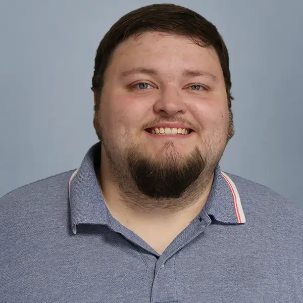 Smiling man with short dark hair and a goatee wearing a blue polo shirt against a light blue background.